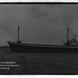 Schwarzweißfoto eines Frachtschiffs im Hafen vor Kranreihe; Logo des Internationalen Maritimen Museums Hamburg links unten