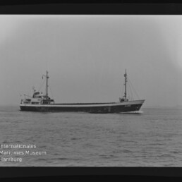 Schwarzweißaufnahme eines schmalen Frachtschiffs mit zwei Masten auf ruhiger See; Schriftzug Internationales Maritimes Museum Hamburg sichtbar.