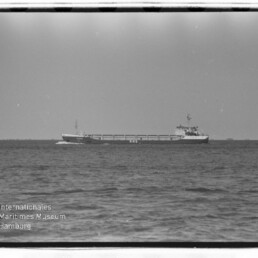 Schwarzweißaufnahme eines Frachtschiffs auf ruhiger See am Horizont, mit Museumswasserzeichen unten links.