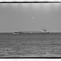 Frachtschiff auf ruhiger See am Horizont, Schwarzweißaufnahme mit Museums-Wasserzeichen unten links.
