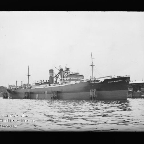 Großes Frachtschiff am Kai mit sichtbaren Aufbauten, Masten und ruhiger Wasserfläche im Vordergrund