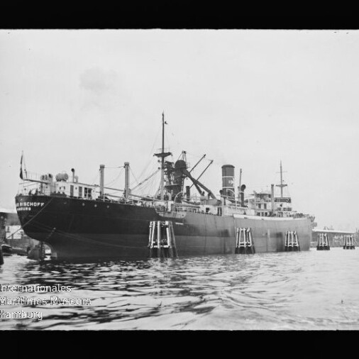 Großes historisches Dampfschiff mit Ladekränen und Schornstein längs eines Kaipiers, Spiegelung im Wasser.