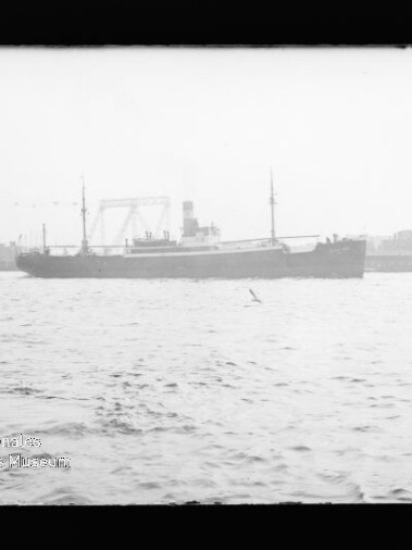 Frachtschiff mit Schornstein auf ruhiger See vor Hafenkränen im Hintergrund, Museumsbeschriftung unten links.