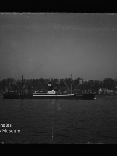 Schwarzweißfoto eines Frachtschiffs auf ruhigem Wasser vor Ufer mit Häusern; Schriftzug 'Maritimes Museum Hamburg' links unten.