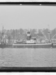 Frachtschiff vor Küste mit Häusern und Bäumen im Hintergrund, Schwarzweißaufnahme mit Museumslogo unten links.