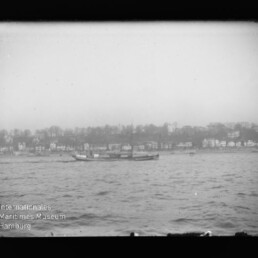 Schwarzweißaufnahme eines niedrigen Passagierschiffs auf rauer See vor einer Uferlinie mit Häusern; Museumslogo unten links.