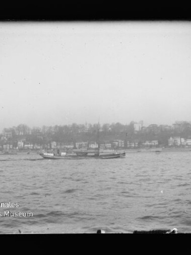 Schwarzweißaufnahme eines niedrigen Passagierschiffs auf rauer See vor einer Uferlinie mit Häusern; Museumslogo unten links.