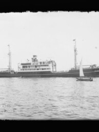 Langes Passagierschiff mit Aufbauten und Schornstein auf ruhigem Wasser, kleines Segelboot im Vordergrund.
