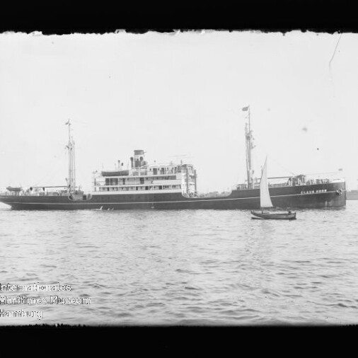 Langes Passagierschiff mit Aufbauten und Schornstein auf ruhigem Wasser, kleines Segelboot im Vordergrund.