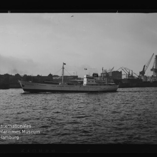 Weißes Frachtschiff auf ruhigem Wasser vor Hafen mit Kränen; schwarzweißes Foto mit Museumswasserzeichen.