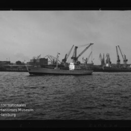Weißes Frachtschiff fährt im Hafen vor Werftkränen; Schwarzweißaufnahme mit Museumslogo links unten.
