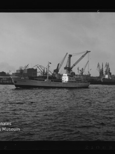 Weißes Frachtschiff fährt im Hafen vor Werftkränen; Schwarzweißaufnahme mit Museumslogo links unten.