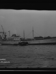 Großes weißes Frachtschiff mit Schlepper im Hafen, Kai mit Kränen im Hintergrund und Wasserzeichen Internationales Maritimes Museum Hamburg.