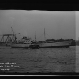 Großes weißes Frachtschiff mit Schlepper im Hafen, Kai mit Kränen im Hintergrund und Wasserzeichen Internationales Maritimes Museum Hamburg.
