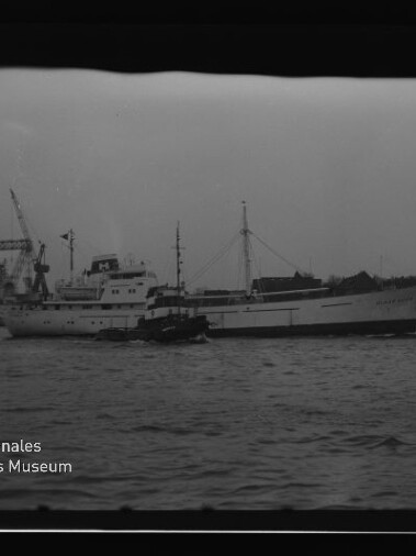 Großes weißes Frachtschiff mit Schlepper im Hafen, Kai mit Kränen im Hintergrund und Wasserzeichen Internationales Maritimes Museum Hamburg.