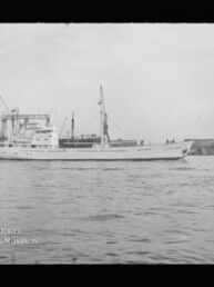 Weißes Frachtschiff mit Mast und Aufbau fährt vor einem Industriehafen mit Kränen, Wellen im Vordergrund.