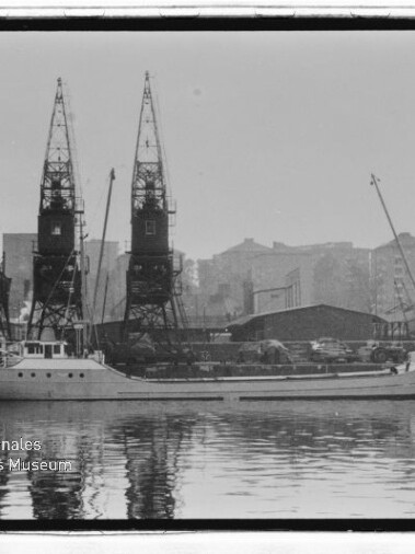 Frachtschiff am Kai vor drei hohen Hafenkränen und Lagerhallen, Spiegelung im Wasser; Museumsschriftzug unten links