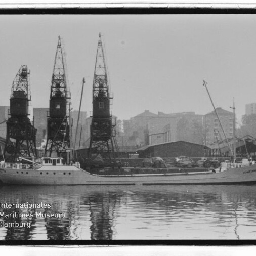 Frachtschiff am Kai vor drei hohen Hafenkränen und Lagerhallen, Spiegelung im Wasser; Museumsschriftzug unten links