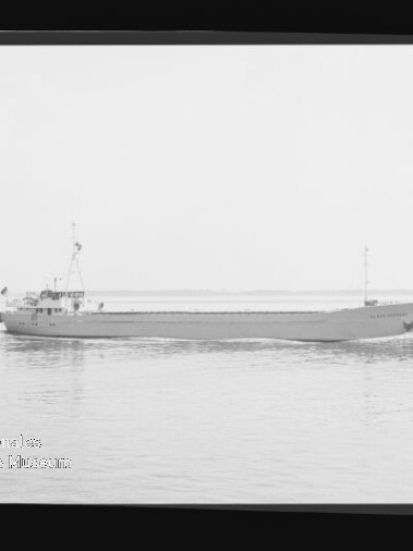 Seitliche Aufnahme eines Frachtschiffs auf ruhigem Wasser in Schwarzweiß, kleines Beiboot am Horizont und Museums-Wasserzeichen links unten.
