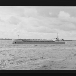 Langes, niedriges Frachtschiff auf ruhiger See unter bewölktem Himmel, mit Horizontlinie und Museumswasserzeichen unten links.