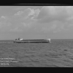 Schwarzweiß-Foto eines länglichen Frachtschiffs auf offener See unter wolkigem Himmel, mit Museumswasserzeichen links unten.
