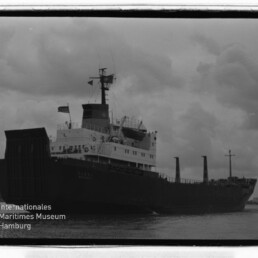 Schwarzweißfoto eines Frachtschiffs im Hafen bei bewölktem Himmel; Wasserzeichen Internationales Maritimes Museum Hamburg