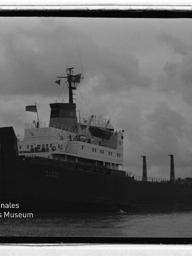 Schwarzweißfoto eines Frachtschiffs im Hafen bei bewölktem Himmel; Wasserzeichen Internationales Maritimes Museum Hamburg