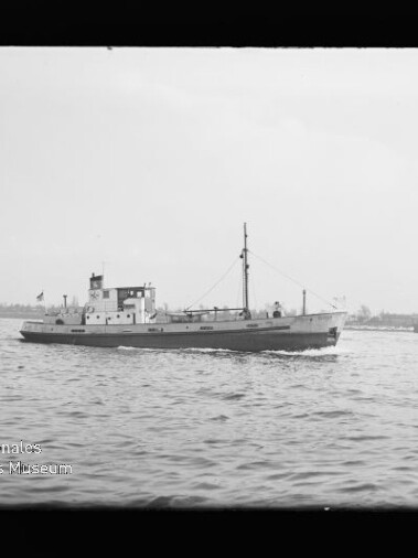 Kleines Versorgungsschiff mit Schornstein und Mast fährt über ruhige See vor flachem Horizont; Museumssignet unten links.