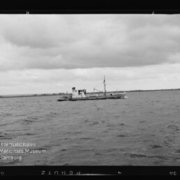 Kleines Schiff mit Mast und Aufbau auf offener See unter bewölktem Himmel, flache Küstenlinie am Horizont.