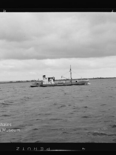 Kleines Schiff mit Mast und Aufbau auf offener See unter bewölktem Himmel, flache Küstenlinie am Horizont.