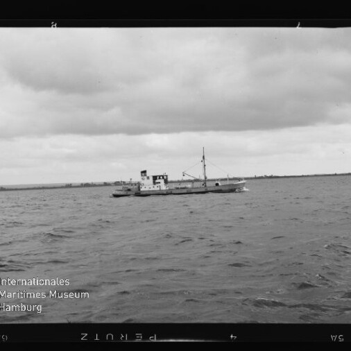 Kleines Schiff mit Mast und Aufbau auf offener See unter bewölktem Himmel, flache Küstenlinie am Horizont.