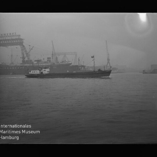 Frachtschiff fährt im Hafen vor Werftkränen in nebliger Stimmung; unten links Schriftzug Internationales Maritimes Museum Hamburg