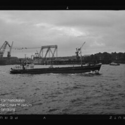Frachtschiff auf bewegter See vor Industriehafen mit Kränen und Hubbrücke, Schriftzug 'Internationales Maritimes Museum Hamburg'.