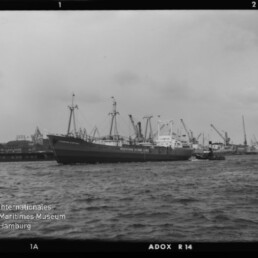 Großes Frachtschiff neben einem Schlepper vor einem Hafen mit zahlreichen Kranen, Schwarzweißaufnahme.