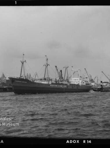 Großes Frachtschiff neben einem Schlepper vor einem Hafen mit zahlreichen Kranen, Schwarzweißaufnahme.