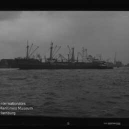 Großes Frachtschiff am Hafen mit Kränen im Hintergrund und Wasserfläche; links unten Logo des Internationalen Maritimen Museums Hamburg
