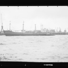 Frachtschiff am Industriehafen mit mehreren Hafenkränen im Hintergrund und aufgewühltem Wasser im Vordergrund.