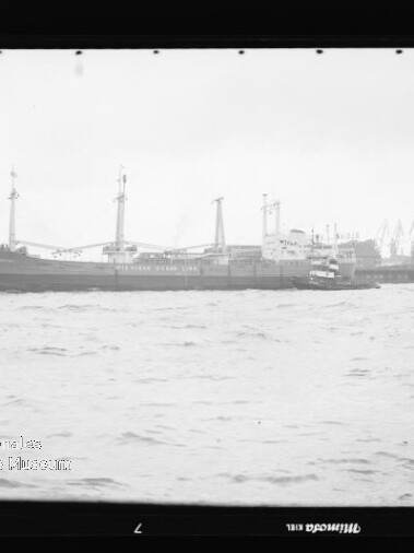 Frachtschiff am Industriehafen mit mehreren Hafenkränen im Hintergrund und aufgewühltem Wasser im Vordergrund.
