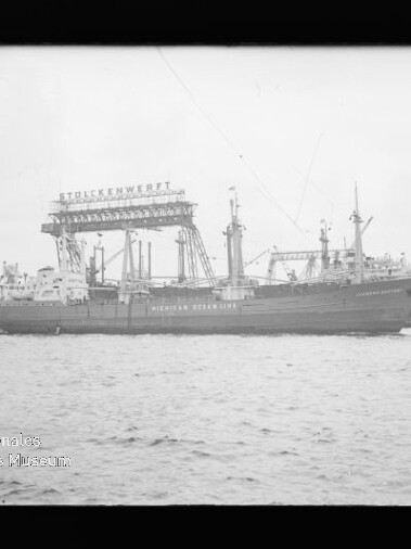 Frachtschiff im Hafen vor einem Werftkran mit Beschriftung Stülckenwerft, Wasser im Vordergrund.