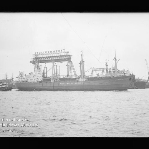 Frachtschiff im Hafen vor einem Werftkran mit Beschriftung Stülckenwerft, Wasser im Vordergrund.