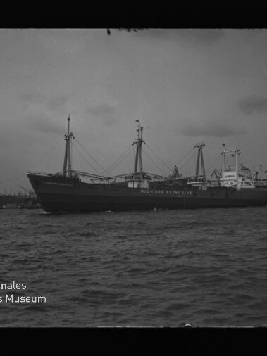Großes Frachtschiff mit Ladebäumen und Masten im Hafen, Wasser im Vordergrund und Kräne im Hintergrund.