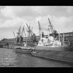Schwarzweißaufnahme eines Frachtschiffs am Kai vor Lagerhallen, mehrere Ladekräne und kleinere Boote im Hafen.