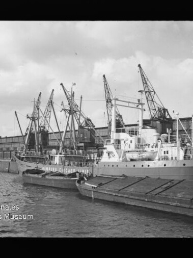 Schwarzweißaufnahme eines Frachtschiffs am Kai vor Lagerhallen, mehrere Ladekräne und kleinere Boote im Hafen.