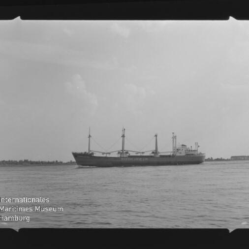 Schwarzweißaufnahme eines Frachtschiffs mit Ladebäumen auf ruhiger See, flache Küstenlinie im Hintergrund sowie Filmrand und Museumslogo.