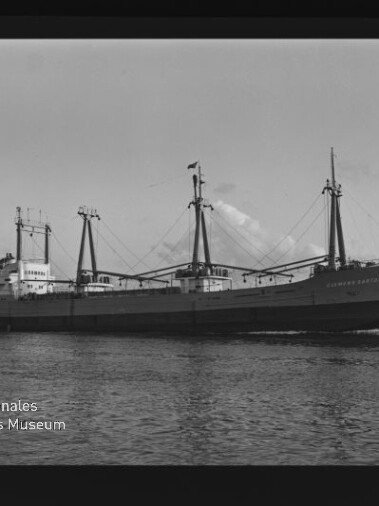 Schwarzweißaufnahme eines Frachtschiffs mit drei Ladebäumen auf ruhigem Meer; Wasserzeichen Internationales Maritimes Museum Hamburg