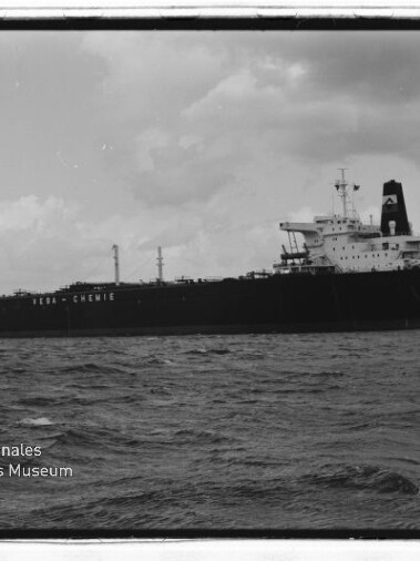 Schwarzweißaufnahme eines großen Tankers mit achterlichem Aufbauten, sichtbarer Bugwelle und bewölktem Himmel