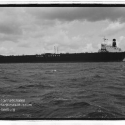 Schwarzweißaufnahme eines großen Tankers mit der Aufschrift VEBA-CHEMIE auf dem Rumpf, ruhige See und wolkiger Himmel.