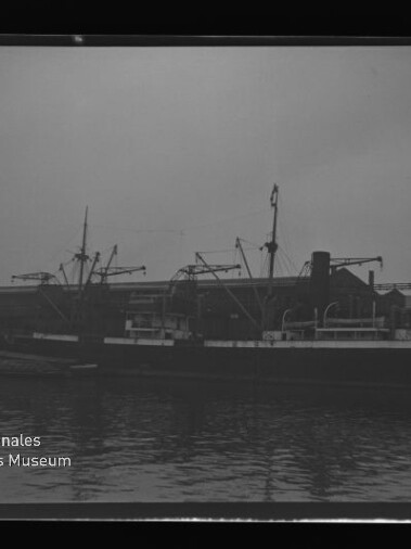 Schwarzweißfoto eines Frachtschiffs am Kai mit Kränen und Lagerhalle; Wasserzeichen Maritimes Museum Hamburg