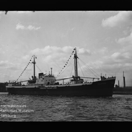 Schwarzweißfoto eines Frachtschiffs mit zwei Masten und Signalfahnen, Aufschrift Hamburg Chicago Line, vor Hafenkulisse.