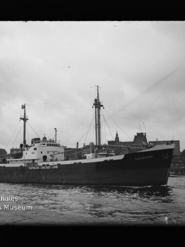 Frachtschiff mit dem Schriftzug Hamburg Chicago Line, Masten sichtbar, auf dem Wasser vor einer Stadtkulisse.